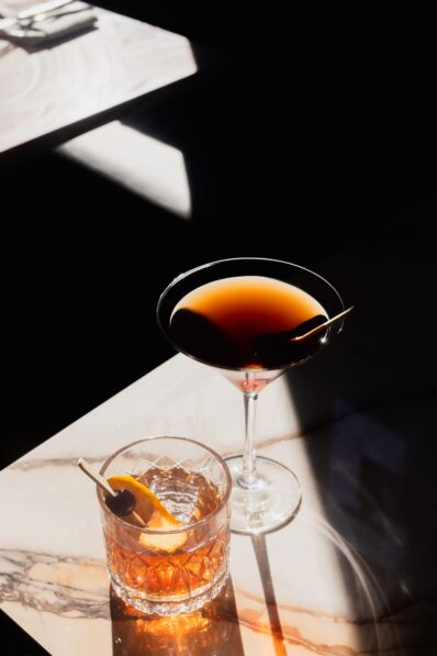 A martini glass with a dark cocktail and a cherry sits beside a tumbler with an orange slice and cherry garnish on a marble table, both drinks highlighted by sunlight and surrounded by deep shadows.