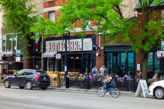 People sit at outdoor tables in front of a bar and restaurant on a city street lined with trees. Cars are parked along the road, and a person rides a bicycle in the foreground.