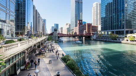People walk along a riverside promenade lined with cafes and trees. Modern glass skyscrapers and a red bridge span the blue-green river under a clear, sunny sky in an urban cityscape.