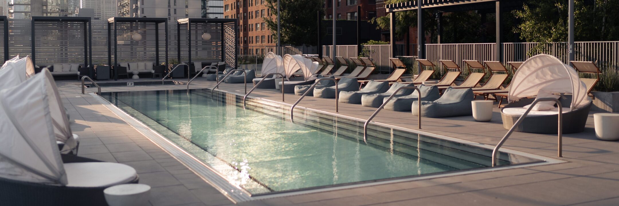 A rooftop pool surrounded by lounge chairs, cabanas, and modern seating, with tall buildings visible in the background on a sunny day.
