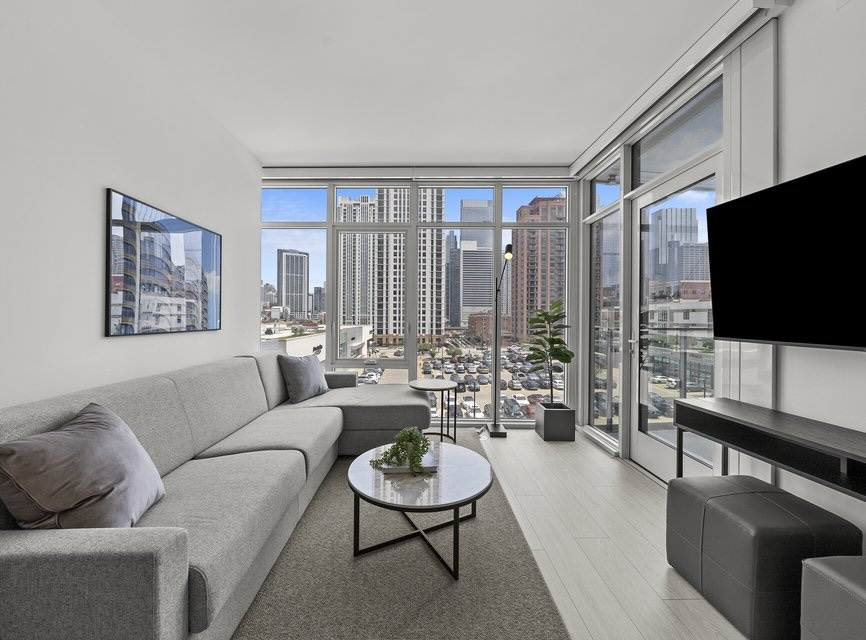 Modern living room with a gray sofa, round coffee table, wall-mounted TV, large windows, and city skyline views. Light walls and flooring create a bright, airy atmosphere. A plant and framed picture add decor.