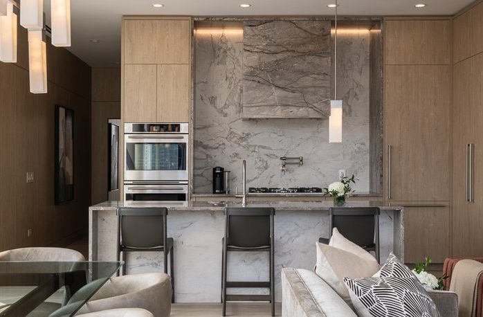 Modern kitchen with light wood cabinets, gray marble backsplash and island, built-in oven, three dark barstools, pendant lights, and neutral-toned furniture in the foreground. A vase with flowers sits on the countertop.