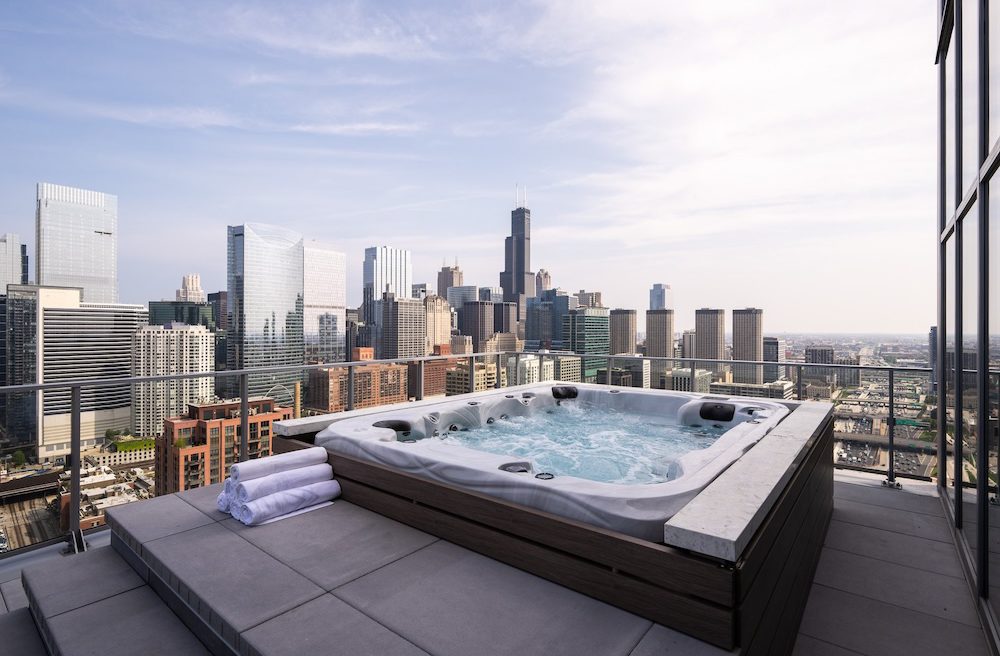 A rooftop hot tub with folded white towels beside it overlooks a modern city skyline with tall buildings under a partly cloudy sky.