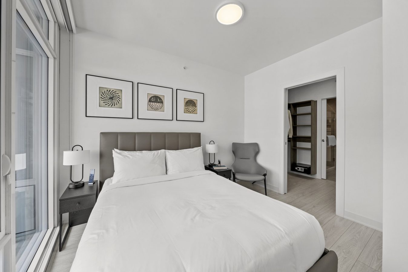 A modern bedroom with a neatly made bed, gray upholstered headboard, nightstands, a gray chair, and wall art above the bed. An open door reveals a closet and bathroom with light-colored flooring.