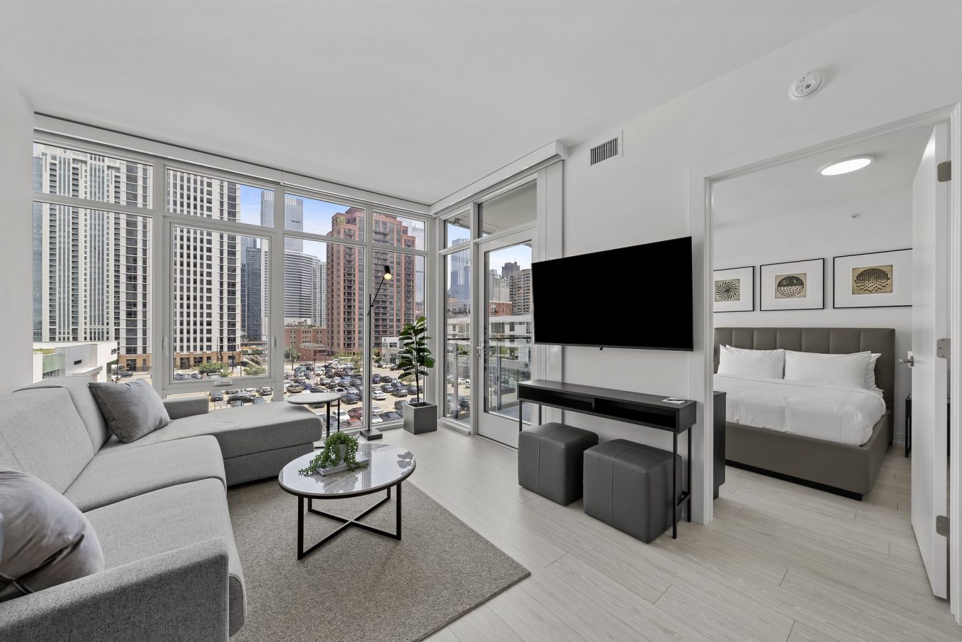 Modern apartment living room with large windows, gray sofa, coffee table, wall-mounted TV, and view of city skyscrapers; bedroom with white bedding visible through an open door.