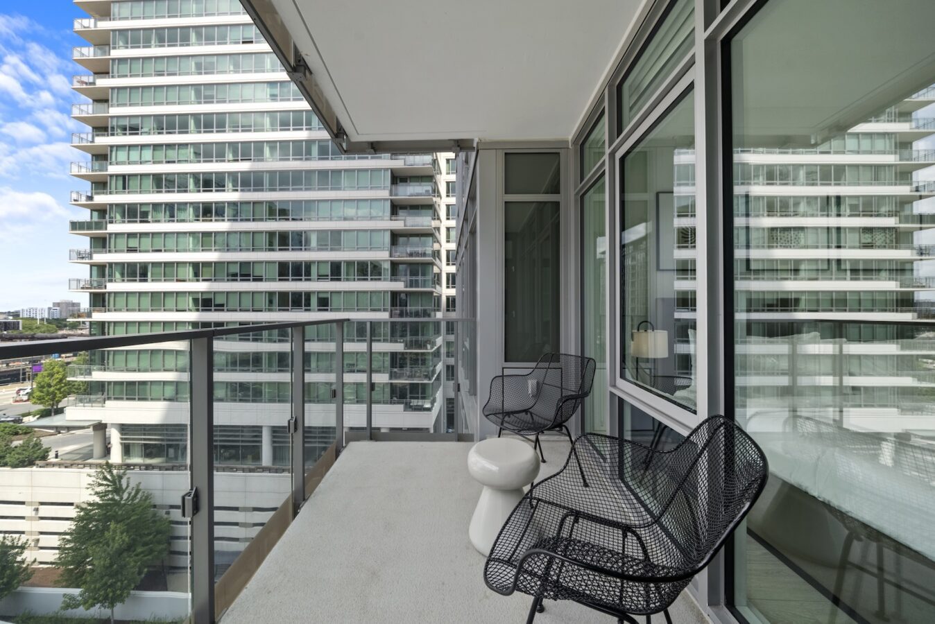 A modern apartment balcony with glass railing, two black wire chairs, and a small white table, overlooking nearby high-rise buildings with many balconies under a blue sky.