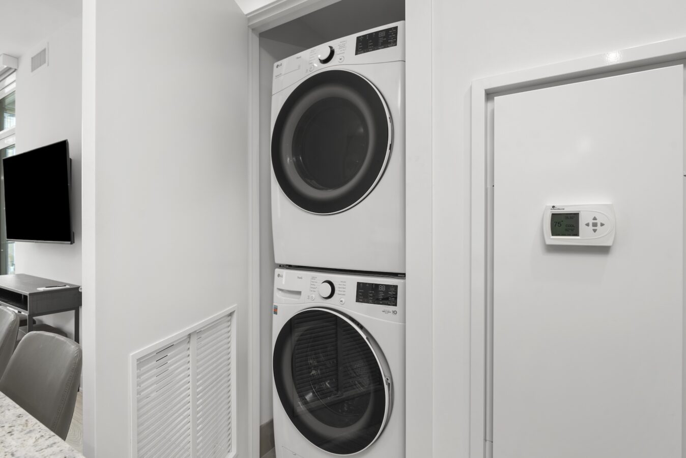 A stacked washer and dryer unit is installed in a white closet next to a wall-mounted thermostat in a modern laundry area with a clean, minimalist design.
