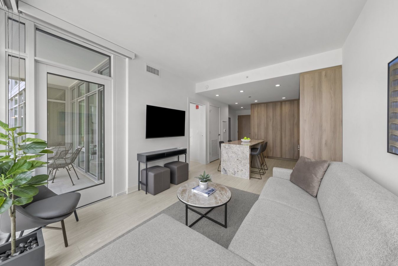 Modern living room with light gray sectional sofa, marble coffee table, wall-mounted TV, and a kitchen area with a central island and wood cabinets. Large glass doors open to a balcony with outdoor seating and potted plants.