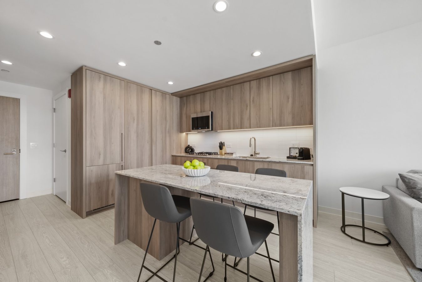 Modern kitchen with light wood cabinets, a marble island with three gray chairs, a bowl of green apples on the island, and built-in appliances. Bright space with wood floors and recessed lighting.