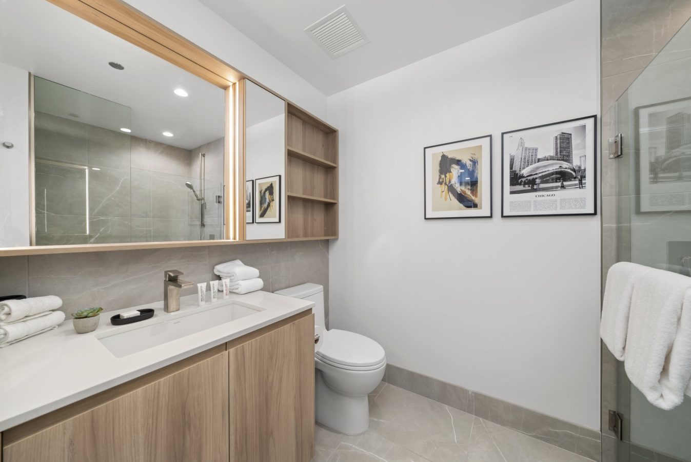 Modern bathroom with light wood cabinets, a white countertop, and a large mirror. There are shelves above the toilet, framed art on the wall, and a glass shower enclosure with a towel hanging on the door.