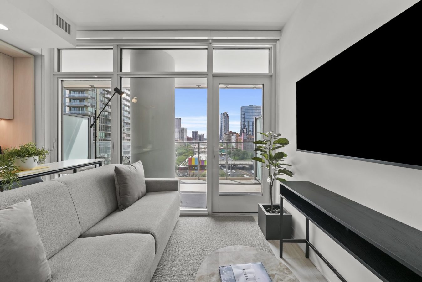 Modern living room with a gray sofa, large TV, potted plant, and floor-to-ceiling windows showing a cityscape with buildings and blue sky. Balcony visible through glass doors.