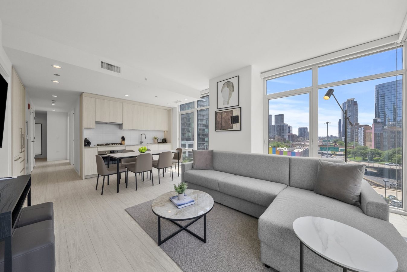 Modern, bright apartment with large windows, a gray sectional sofa, round coffee table, open kitchen and dining area, light wood floors, city skyline visible outside.