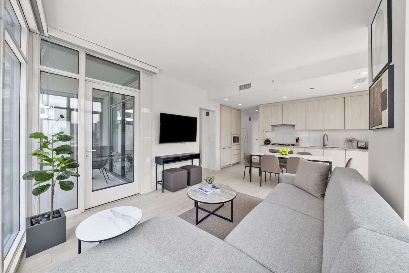 Modern open-concept living room and kitchen with light wood cabinets, gray sectional sofa, wall-mounted TV, small round tables, indoor plant, and a glass door leading to a balcony. Bright, minimalistic decor.