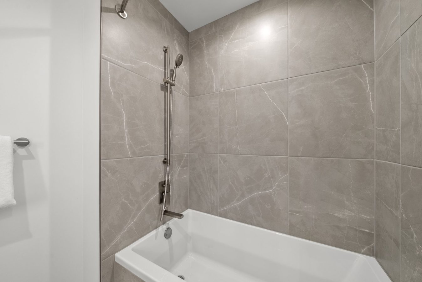 Modern bathroom with large gray marble tiles, a white bathtub, and a wall-mounted showerhead. A folded white towel hangs on a silver hook to the left outside the shower area.