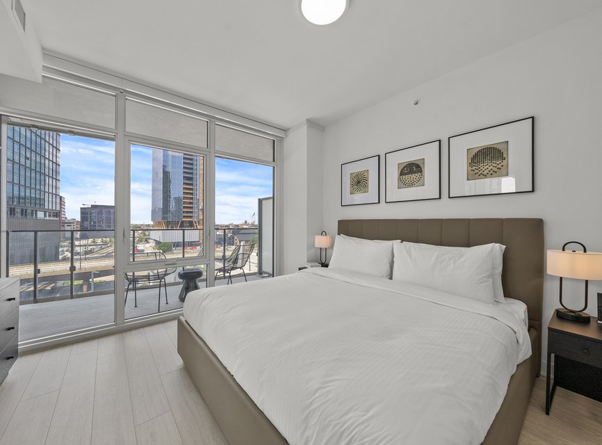 A modern bedroom with a large bed, white linens, bedside tables with lamps, three framed artworks on the wall, and floor-to-ceiling windows opening to a balcony with city views.