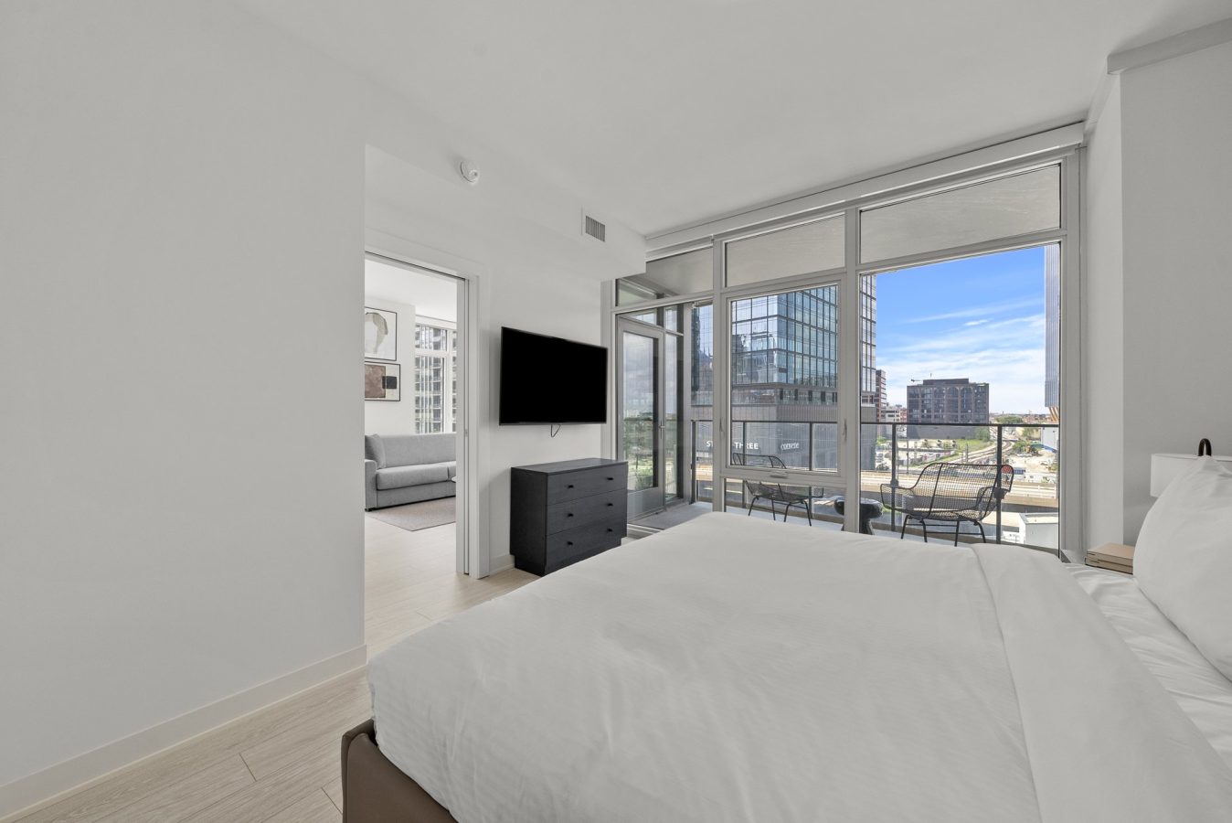 Modern bedroom with white walls, a bed with white linens, a dresser with a wall-mounted TV, and large floor-to-ceiling windows leading to a balcony with outdoor seating overlooking city buildings.