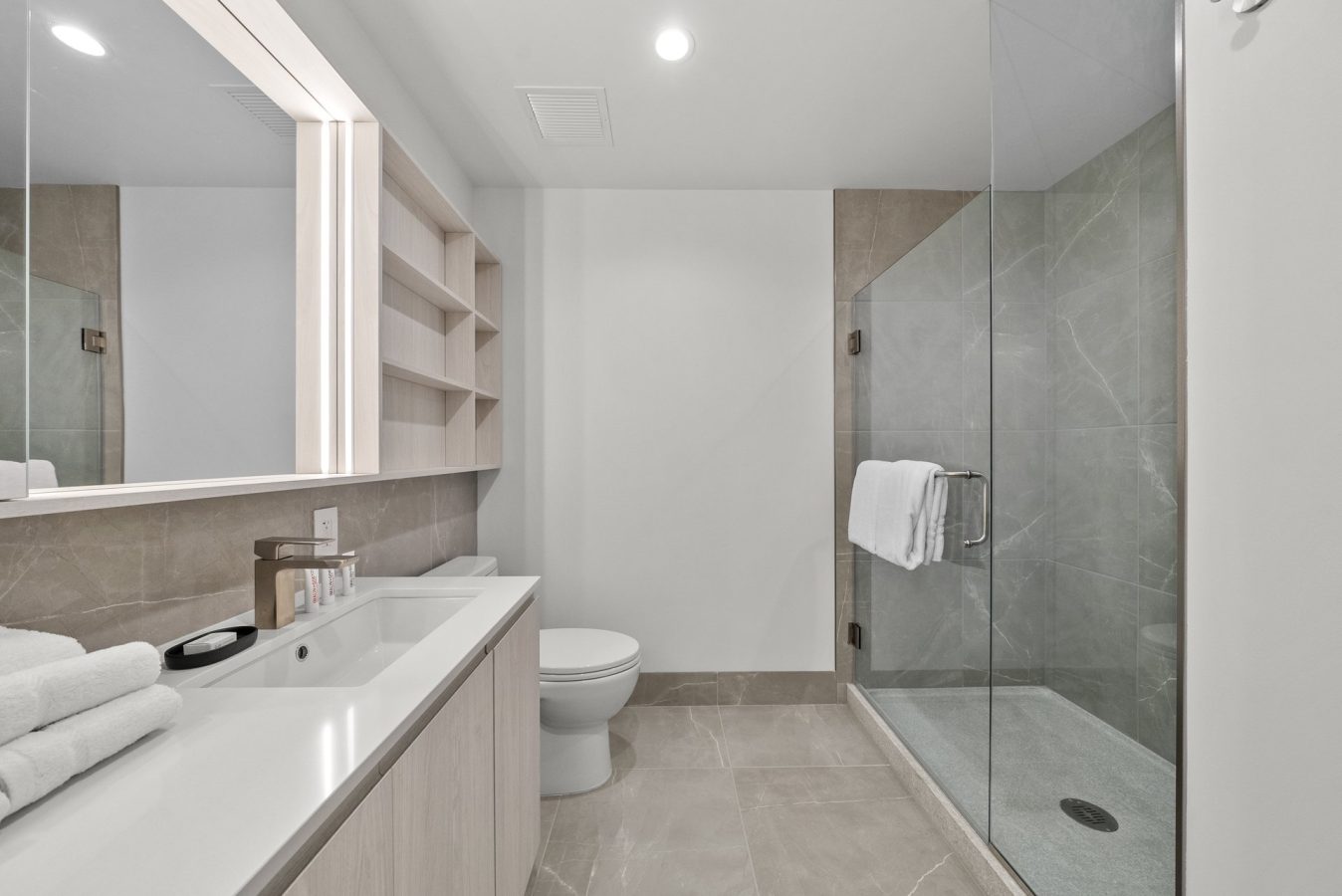 Modern bathroom with light-colored tile flooring, a glass-enclosed shower, toilet, large mirror, built-in shelves, and a vanity with a sink, soap, toiletries, and neatly folded towels.