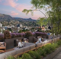 People relax on a rooftop patio with modern seating, greenery, and flowers. The view overlooks a cityscape with hills and the Hollywood sign visible in the distance under a pink sunset sky.