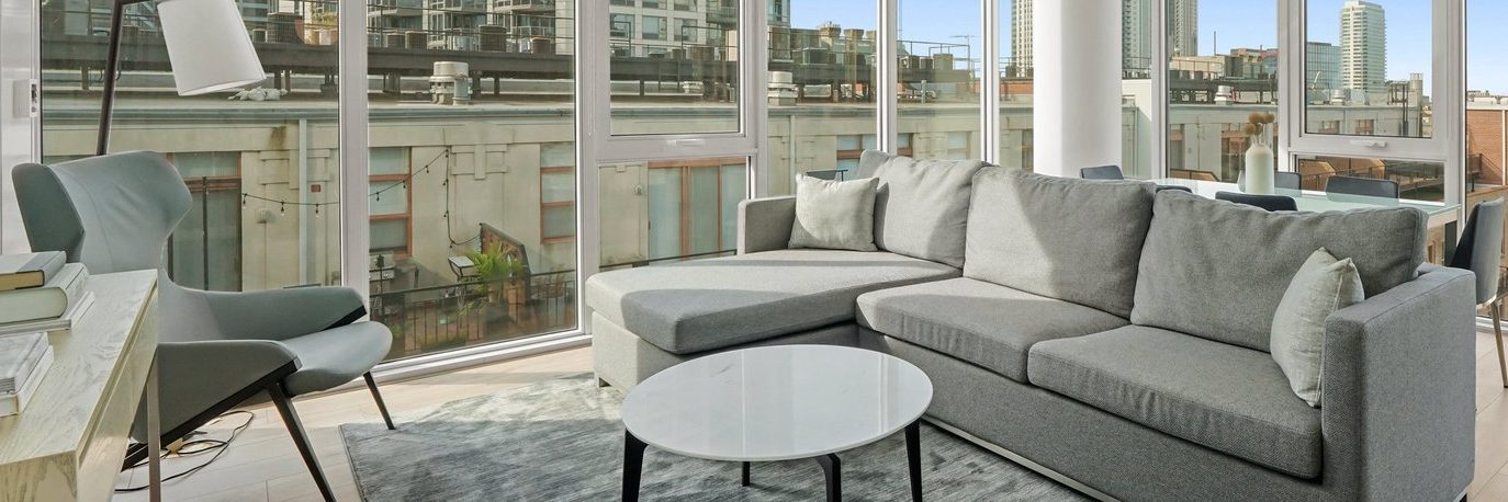 A modern living room with a large gray sectional sofa, round white coffee table, armchair, and floor-to-ceiling windows overlooking city buildings.