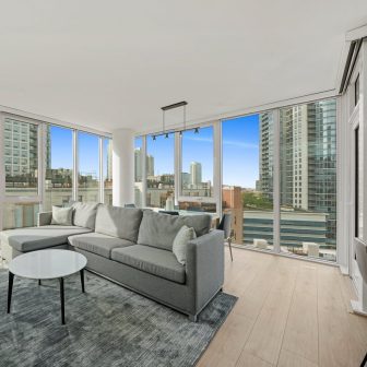 A modern living room with large floor-to-ceiling windows, a gray sectional sofa, a round white coffee table, a gray rug, and city skyscrapers visible outside against a clear blue sky.