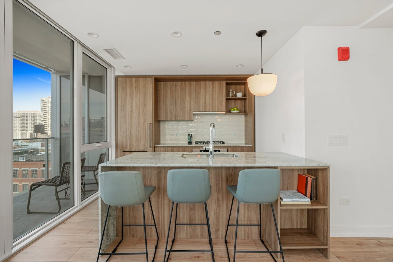 Modern kitchen with wood cabinets, marble island, three blue barstools, and pendant light. Large windows show a city view and a balcony with two chairs. Shelves hold dishes; a book and vase sit on the island.
