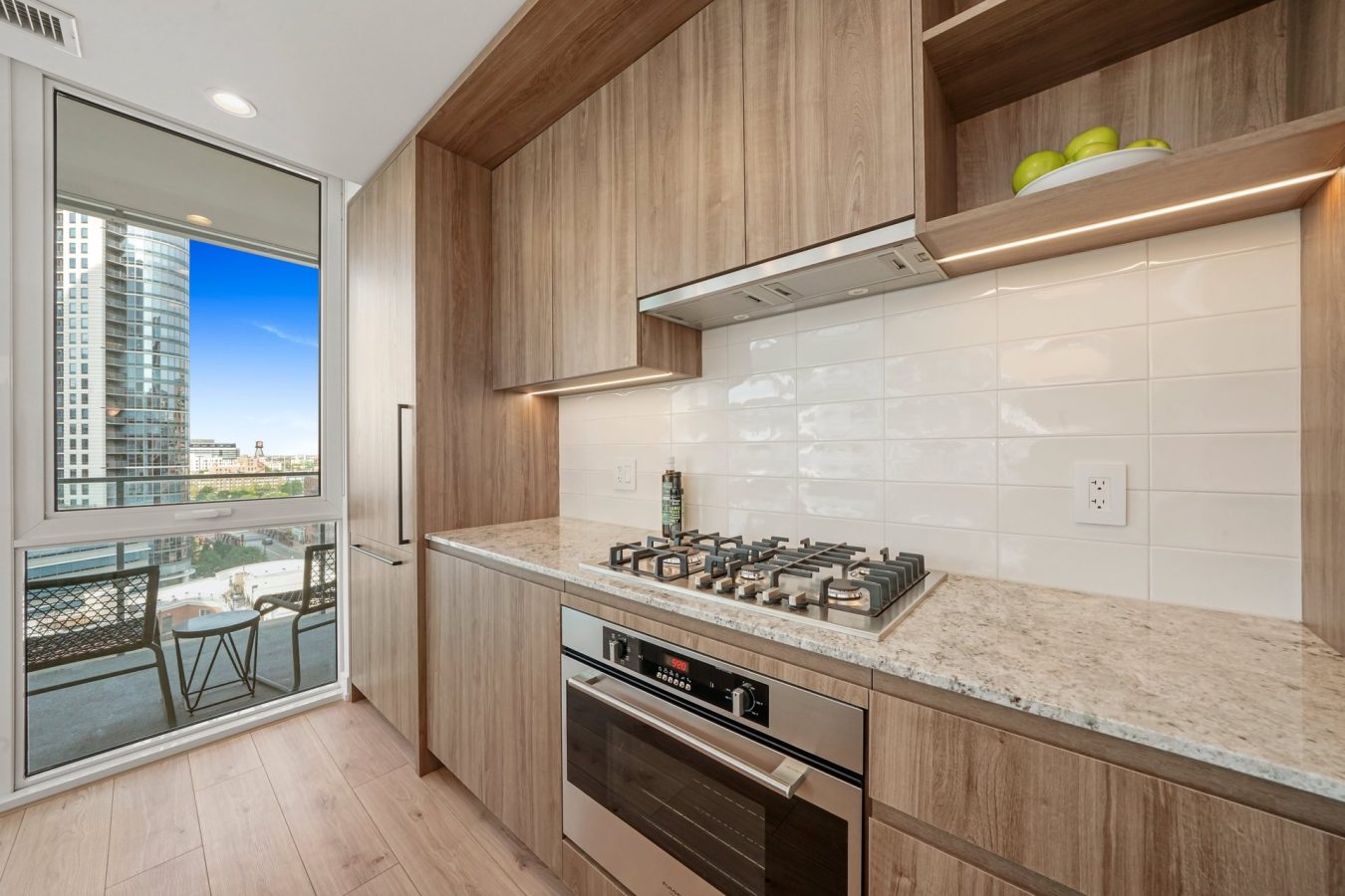 Modern kitchen with wood cabinets, a gas stovetop, built-in oven, white tile backsplash, and light granite countertops. Large window and glass door open to a balcony with city views and outdoor seating.