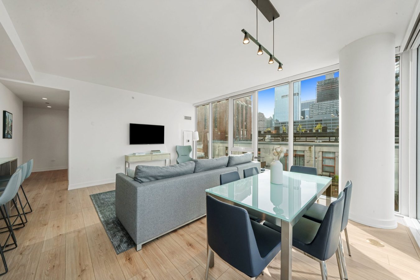 Modern living and dining area with large windows, city views, gray sofa, TV, glass dining table with blue chairs, light wood floors, and bright natural light.