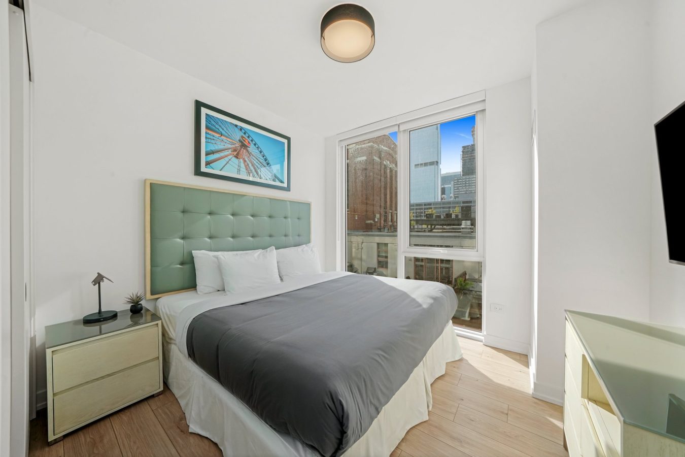 A modern, bright bedroom with a large bed, gray bedding, green tufted headboard, nightstands, wall art, a mounted TV, wood floors, and large windows showing a city skyline view.