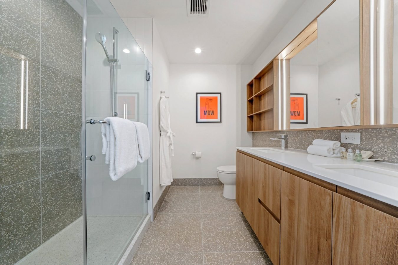 Modern bathroom with a glass-enclosed shower, wooden vanity with double sinks, large mirror, toilet, wall art, and two white robes hanging on the wall. Towels and toiletries are neatly arranged.