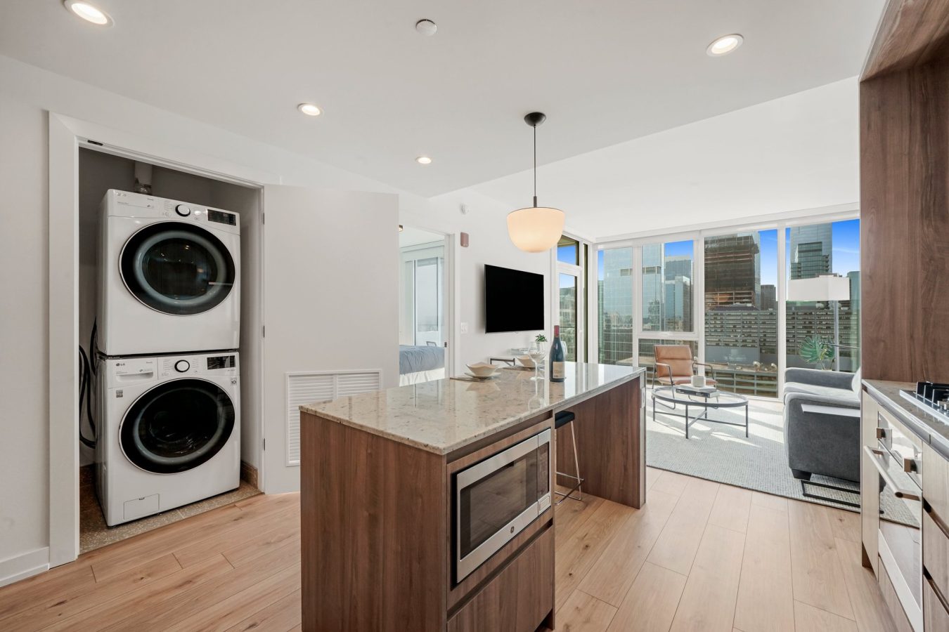 Modern apartment kitchen with wood floors, an island, stacked washer and dryer in an open closet, and large windows offering a city skyline view. Living area has a sofa, TV, and natural light.