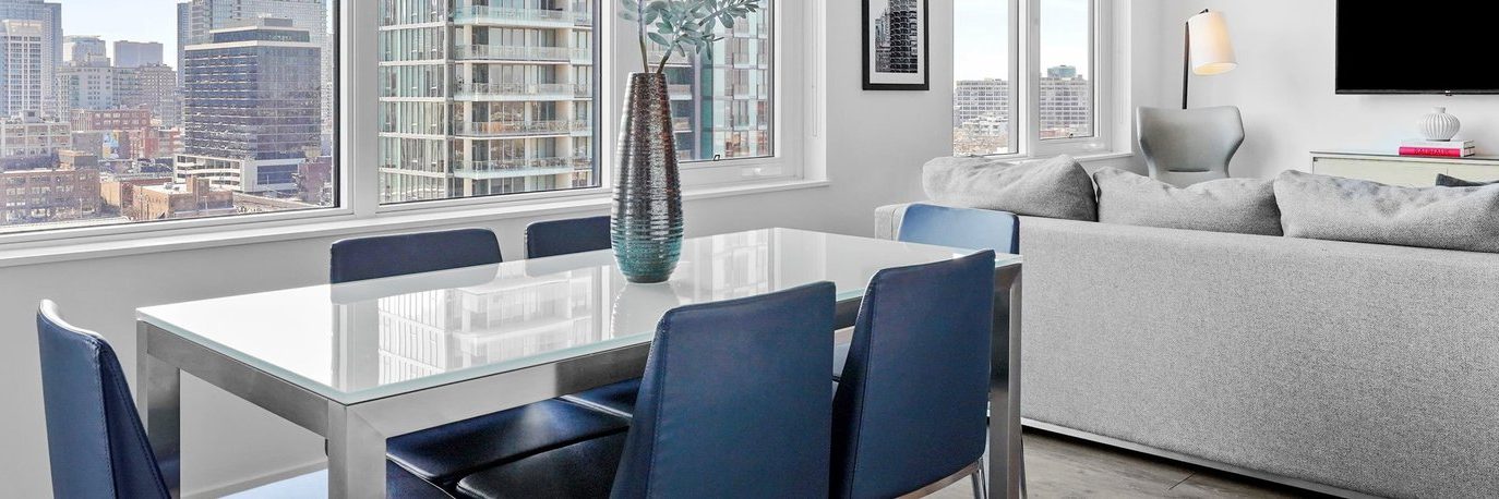 A modern, bright apartment with large windows, a glass dining table with dark chairs, a gray sofa, and a city skyline view in the background. A vase with greenery sits on the table.