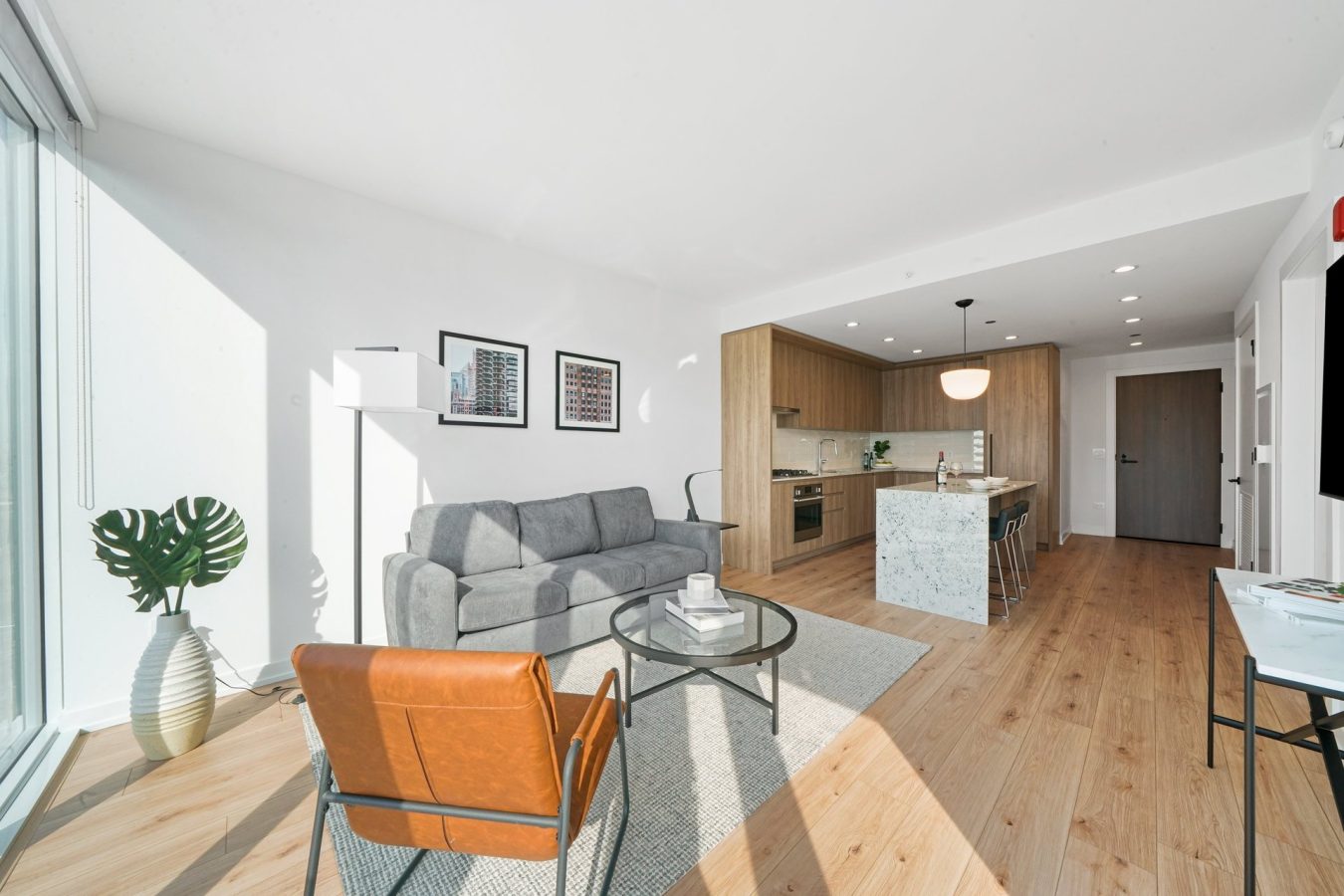 Bright, modern living room with large windows, a gray sofa, brown leather chair, round glass coffee table, and open kitchen featuring wood cabinets and a marble island on light wood floors.