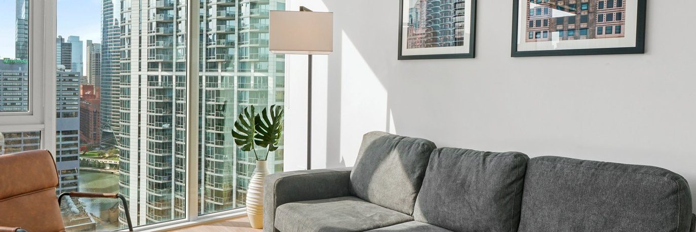 A modern living room with a gray sofa, brown armchair, floor lamp, plant in a vase, and framed city photos on the wall, featuring large windows overlooking city skyscrapers.
