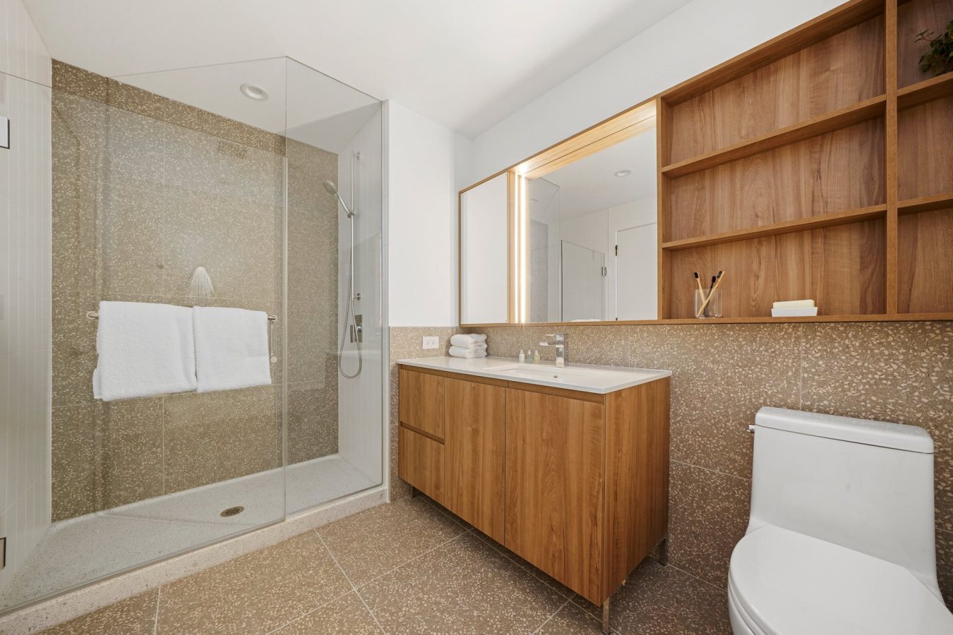 Modern bathroom with a glass-enclosed shower, double towel rack, wood vanity with sink, large mirror, open wooden shelves, and a white toilet, all set against beige textured walls and floor.