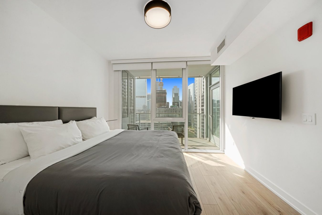 Modern bedroom with a large bed, gray and white bedding, wall-mounted TV, and floor-to-ceiling windows offering a city skyline view. A small balcony can be seen through the glass doors.