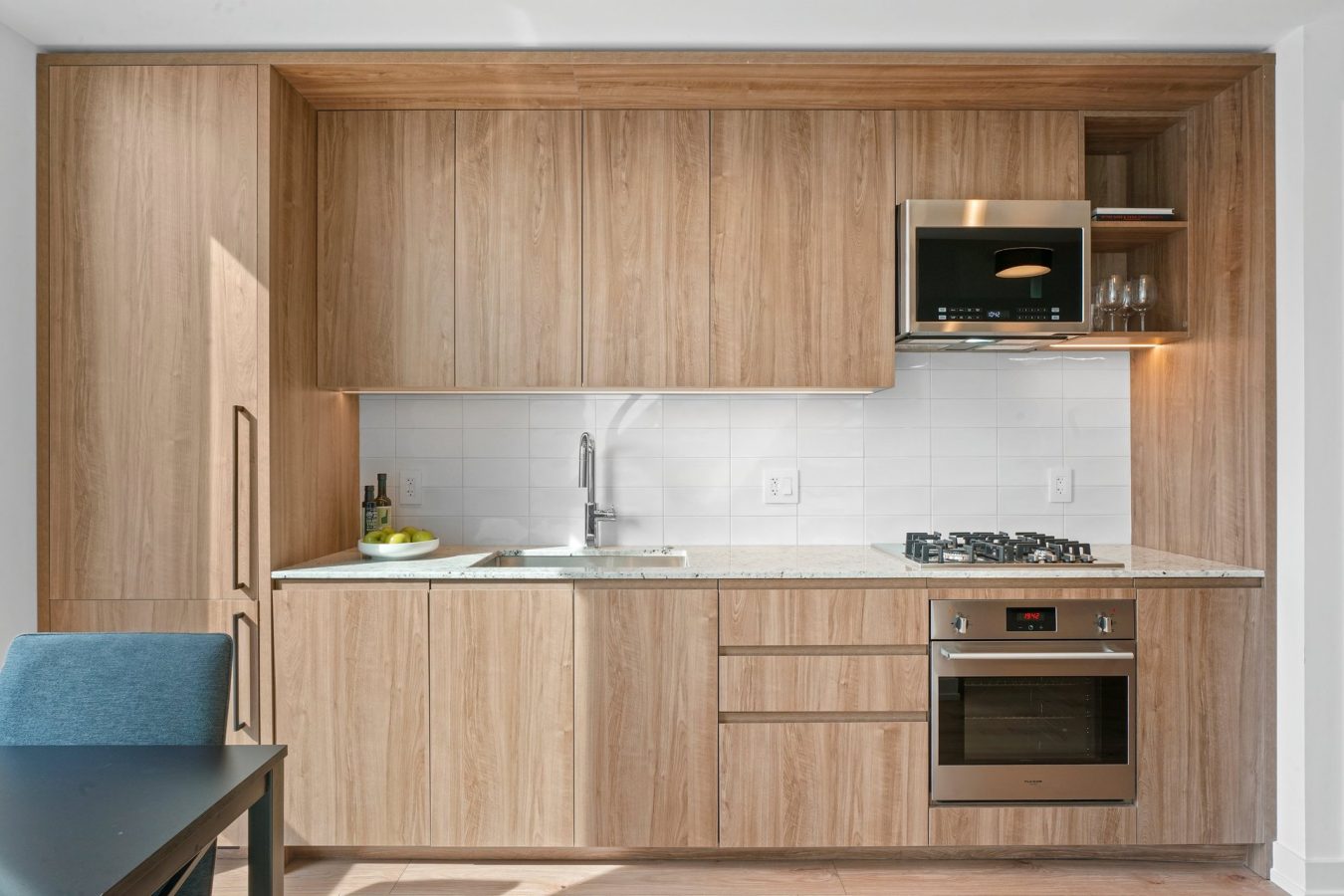Modern kitchen with light wood cabinets, white tile backsplash, stainless steel oven, gas stove, microwave, sink, and a black dining table with a blue chair. A bowl of fruit and bottles are on the counter.