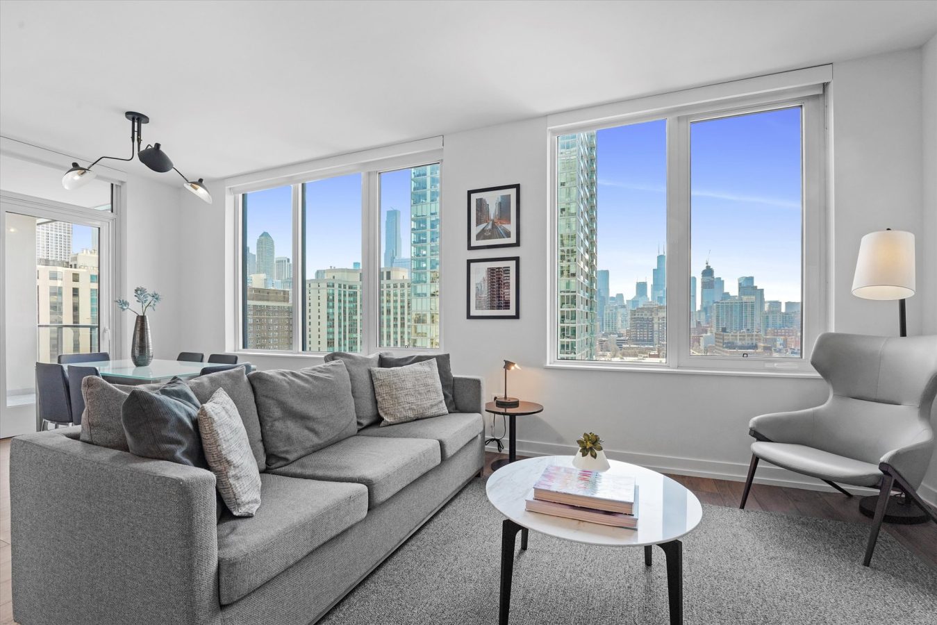 Modern living room with large windows showing a city skyline, gray sofa with cushions, armchair, floor lamp, coffee table with books, and dining area in the background. Bright, contemporary decor with urban views.