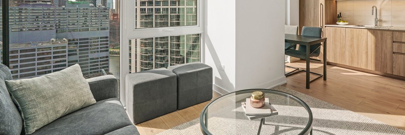 A modern apartment living area with large windows overlooking city skyscrapers, a gray sofa, two ottomans, a round glass coffee table, and a kitchen area with wooden cabinets and a countertop.