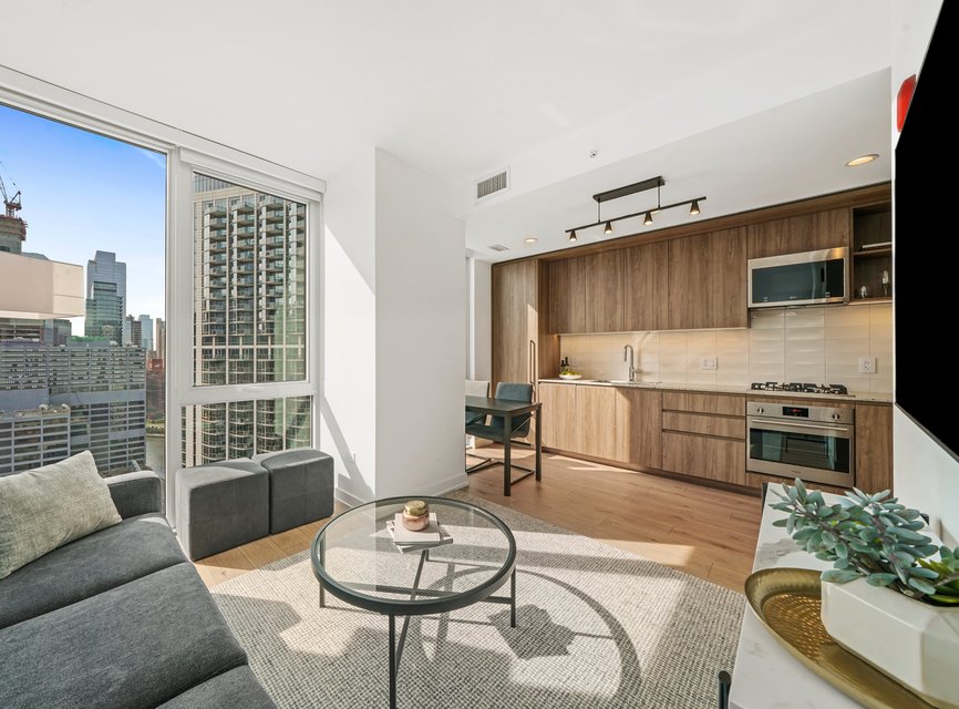 A modern apartment living room with a gray sectional sofa, round glass coffee table, flat-screen TV, and open kitchen featuring wood cabinets and stainless steel appliances. Large windows offer a city skyline view.
