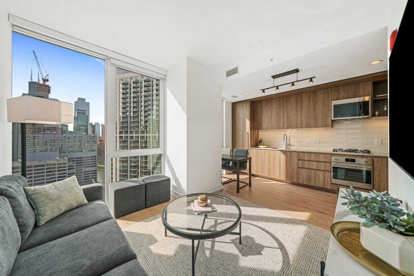 Modern high-rise apartment living room with a gray sofa, round glass coffee table, and open kitchen with wood cabinets. Large windows show a city skyline with tall buildings and construction cranes.