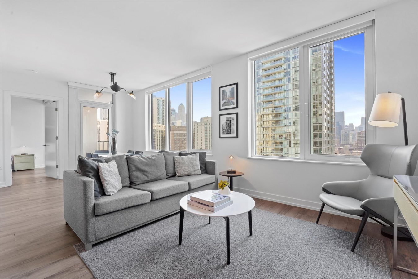 Modern living room with large windows showing a city skyline, gray sofa, round coffee table with books, gray armchair, floor lamp, and framed wall art. Light wood flooring and neutral decor create a bright, airy space.