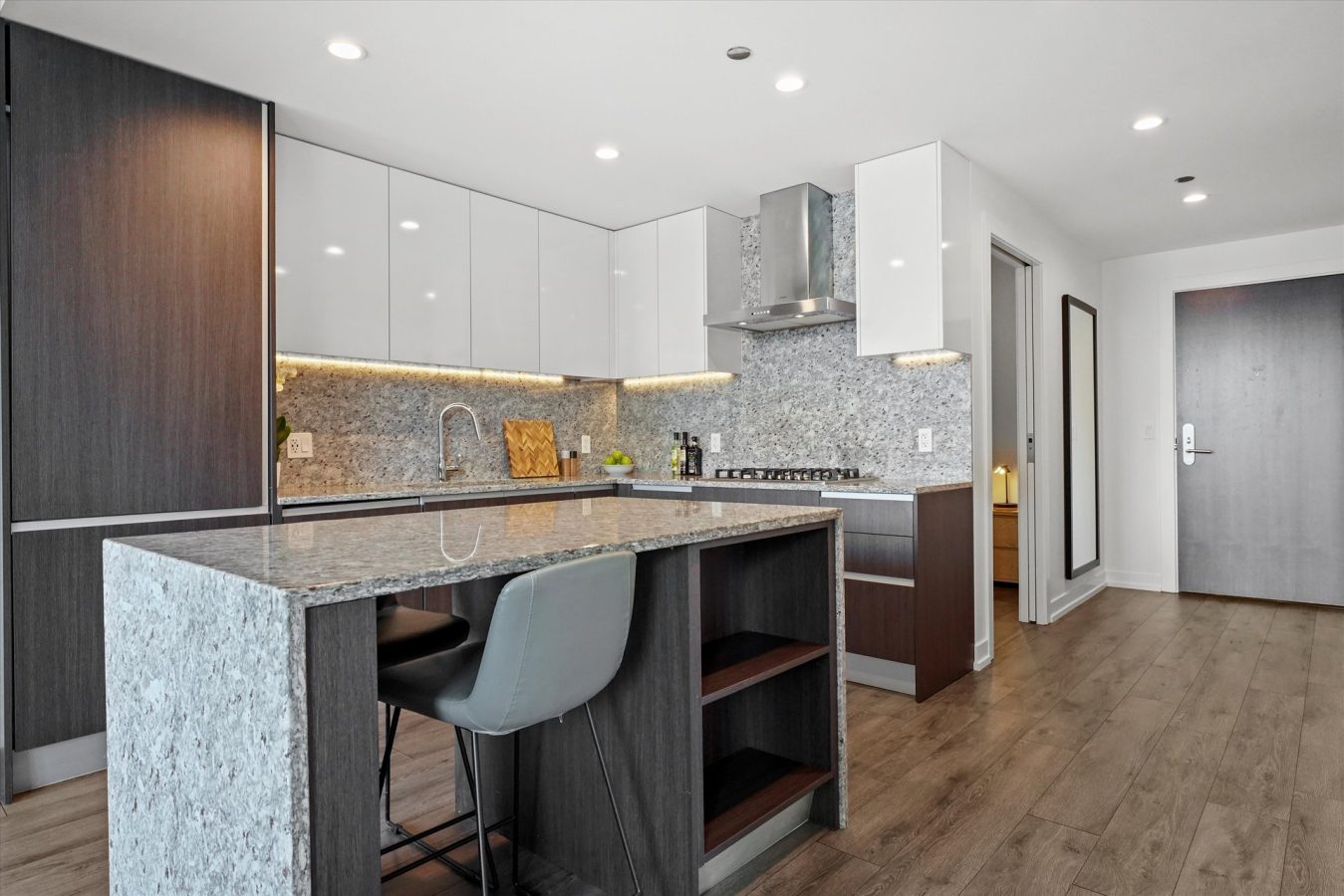 Modern kitchen with dark wood cabinets, white upper cabinets, and gray granite countertops. An island with a barstool is in the foreground, and stainless steel appliances are built into the cabinetry.