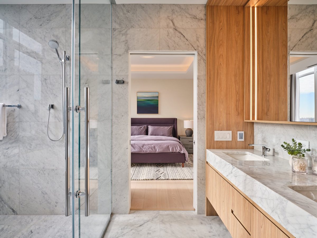 A modern bathroom with marble walls and countertops, a glass-enclosed shower, and wooden cabinets. Through the doorway, a bedroom with a purple bed and a painting above the headboard is visible.