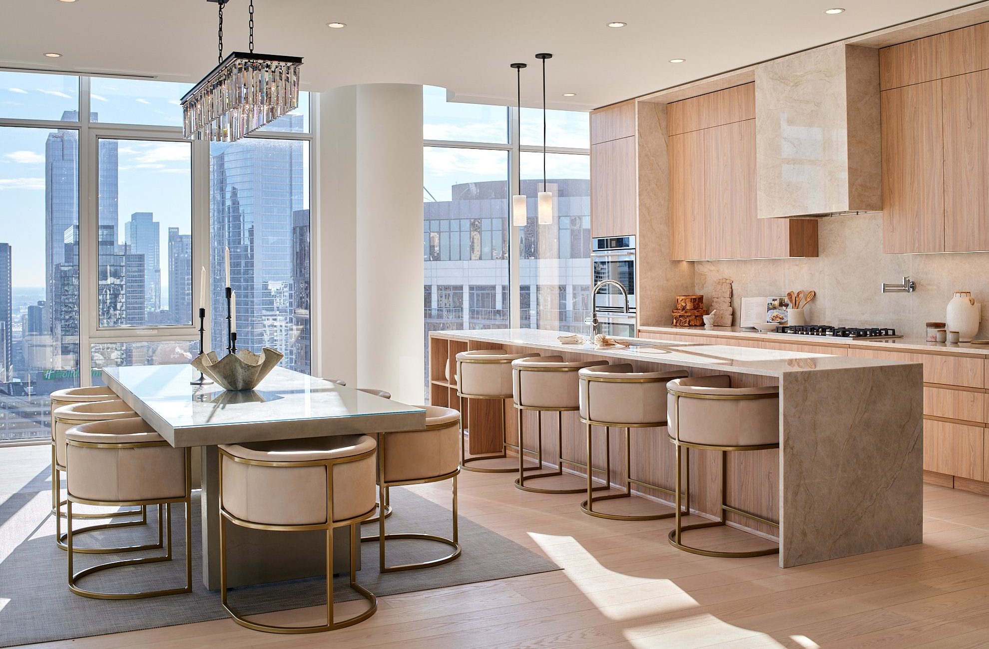 Modern kitchen and dining area with light wood cabinetry, marble countertops, a long island with six beige and gold barstools, a large dining table, floor-to-ceiling windows, and city skyline views.