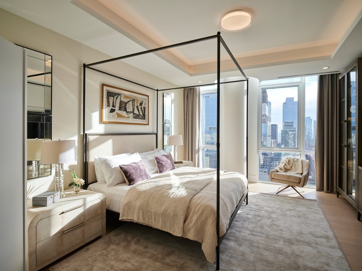 A modern bedroom with a canopy bed, neutral bedding, nightstands with lamps, an abstract painting above the bed, a cozy chair by large windows, and a city skyline view. Natural light fills the space.
