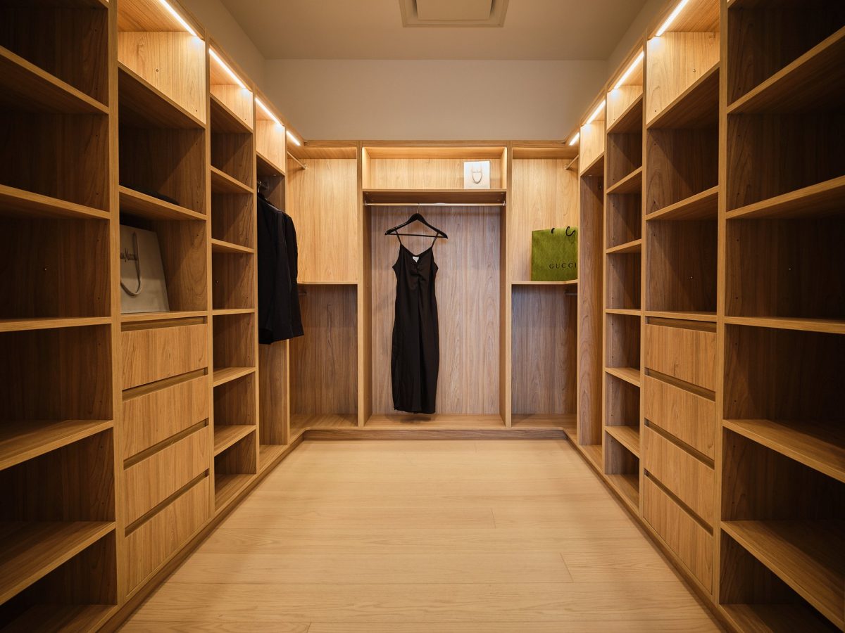 Spacious walk-in closet with wooden shelves and drawers, featuring a black dress and jacket hanging on display. A green shopping bag sits on a shelf. The closet is brightly lit with warm lighting.