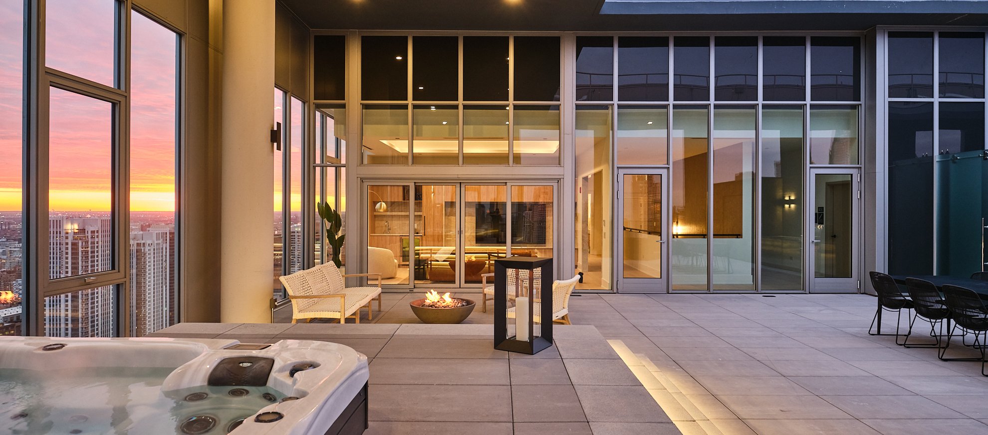 Modern rooftop terrace at sunset with a hot tub, fire pit, and outdoor seating. Large glass doors reveal a stylish indoor lounge. City skyline visible in the background. Warm, inviting lighting throughout.