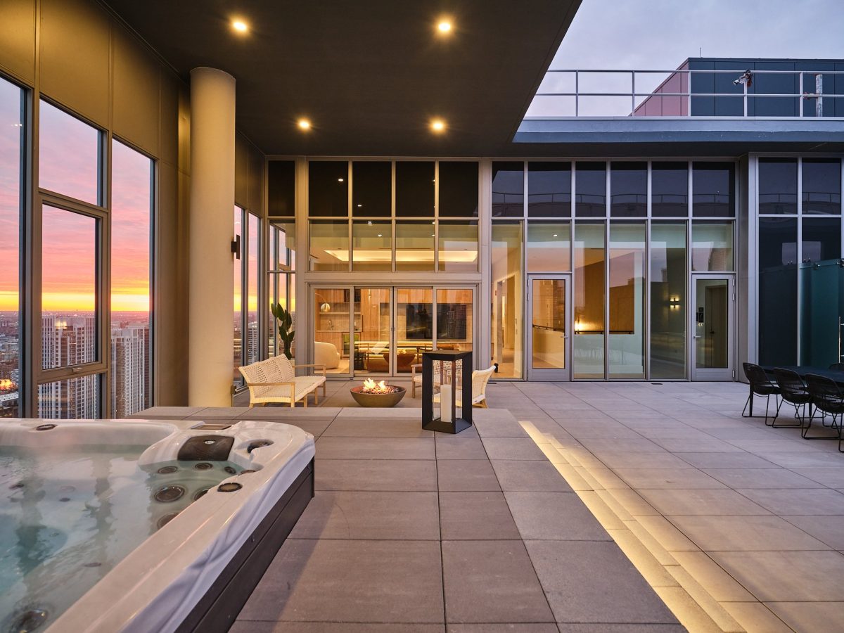 Modern rooftop terrace at sunset with a hot tub, lounge seating, fire pit, and large glass windows overlooking a city skyline. Warm lights illuminate the spacious, stylish outdoor area.