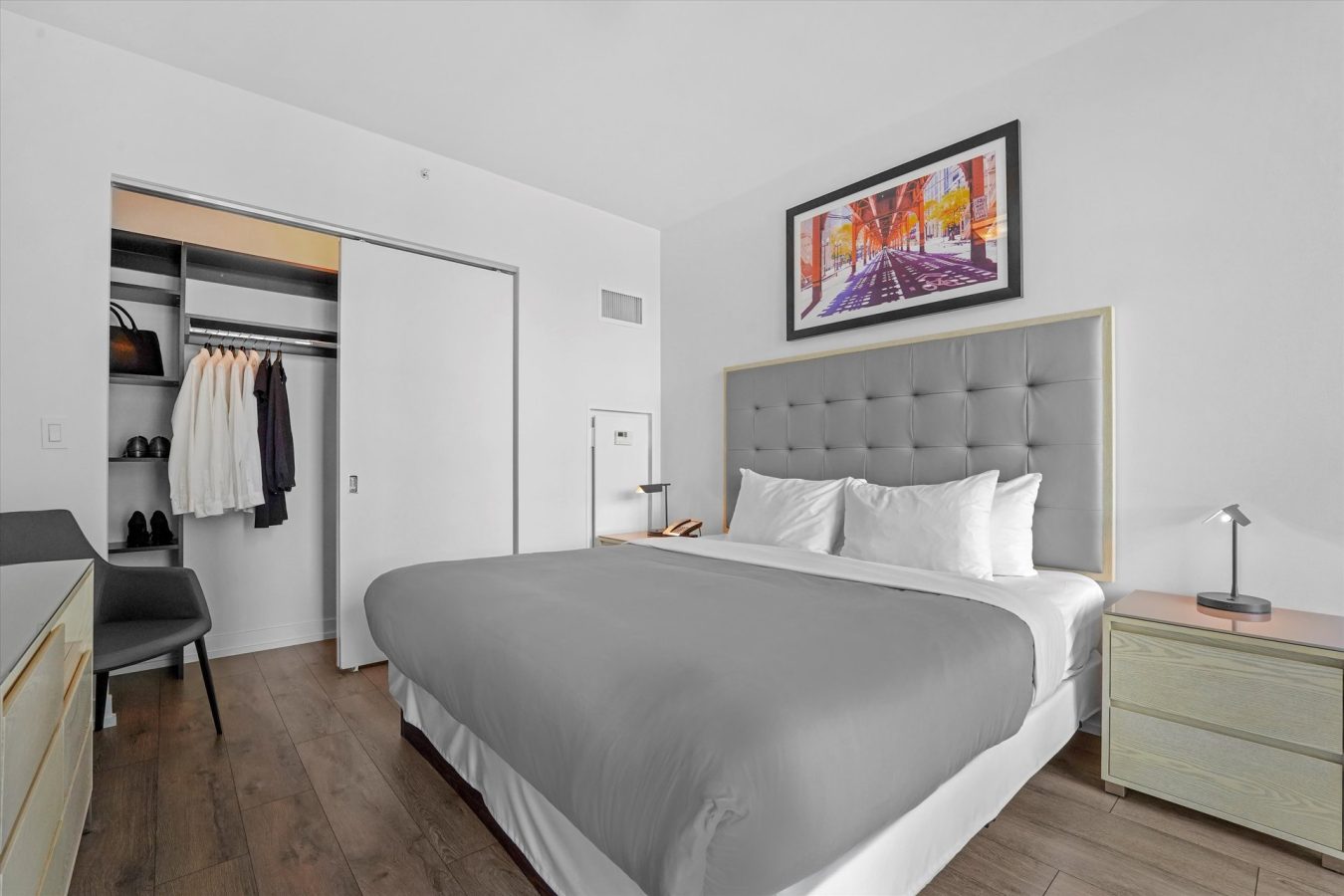 A modern, minimal bedroom with a neatly made bed, gray headboard, nightstands, a painting above the bed, an open closet with clothes and shoes, wood floors, and a desk with a chair.