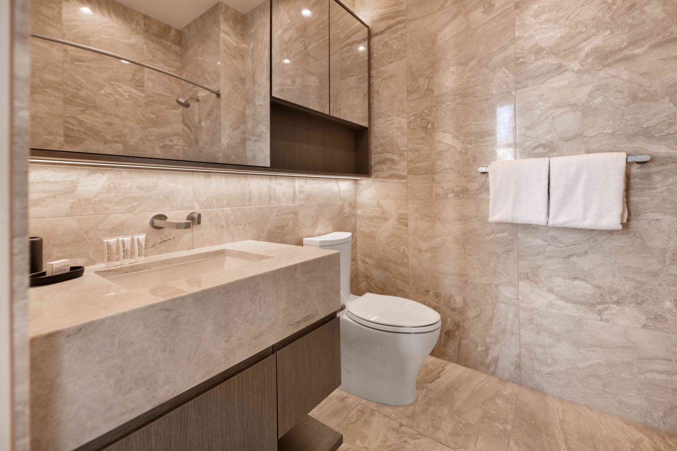 A modern bathroom with beige marble walls and floors, a wall-mounted sink with toiletries, a toilet, a large mirror above the sink, and two folded white towels on a towel rack.